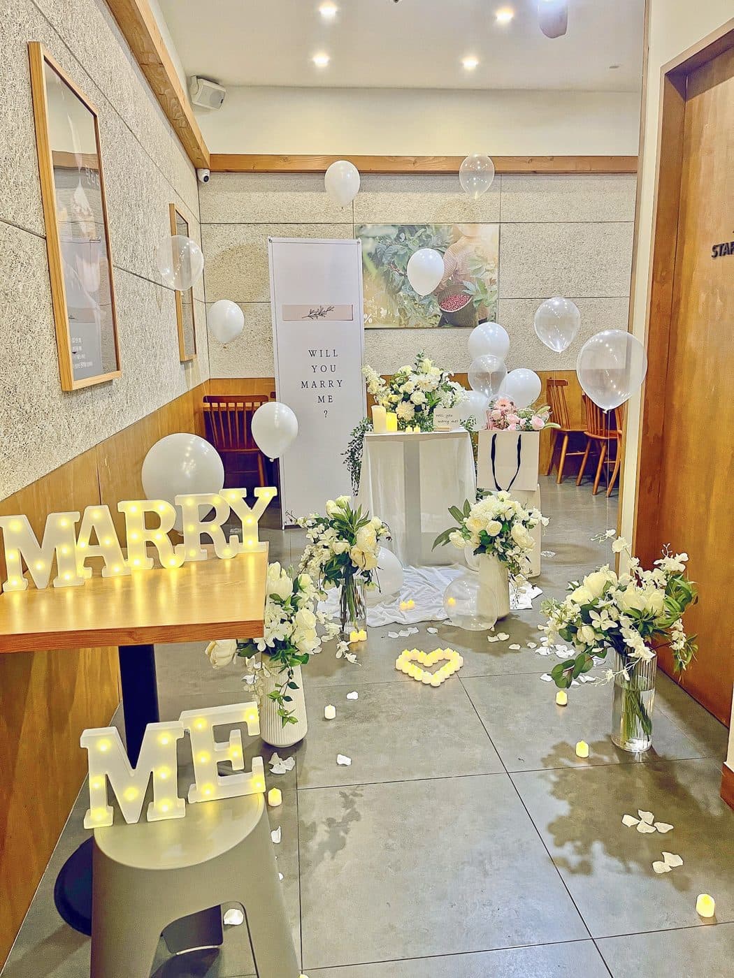 카페 한쪽에 흰 꽃다발 네 개와 MARRY ME 조명을 놓고 캔들로 하트를 만든 프로포즈 현장